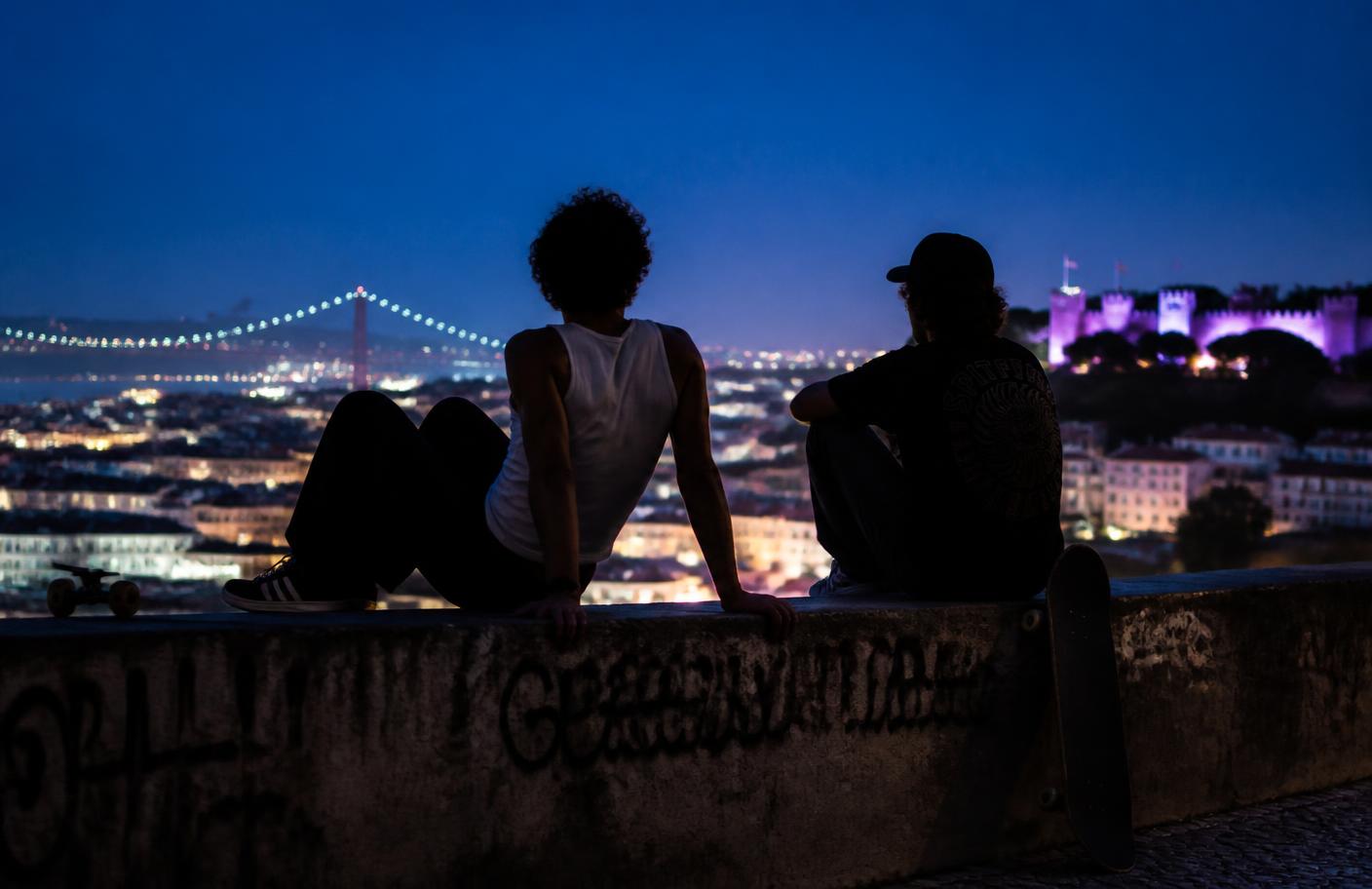 Two skaters watching Lisbon light up from a viewpoint wall